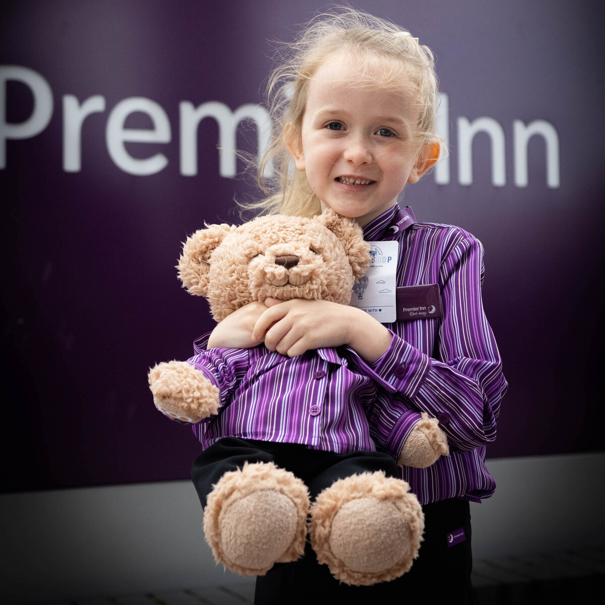 A girl stood in front of a Premier Inn sign wearing a custom made uniform, holding a teddy bear.