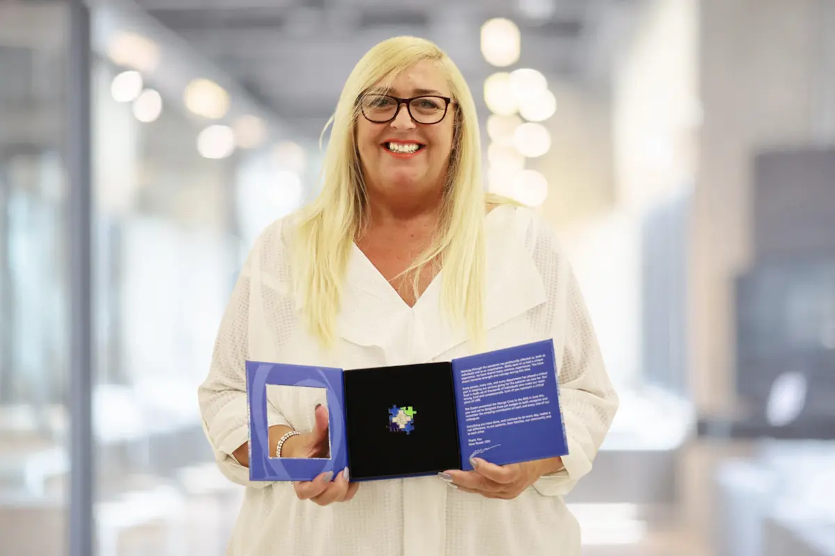 Roma is a blond female stood smiling to camera holding her award, placed in a box, from the NHS. 