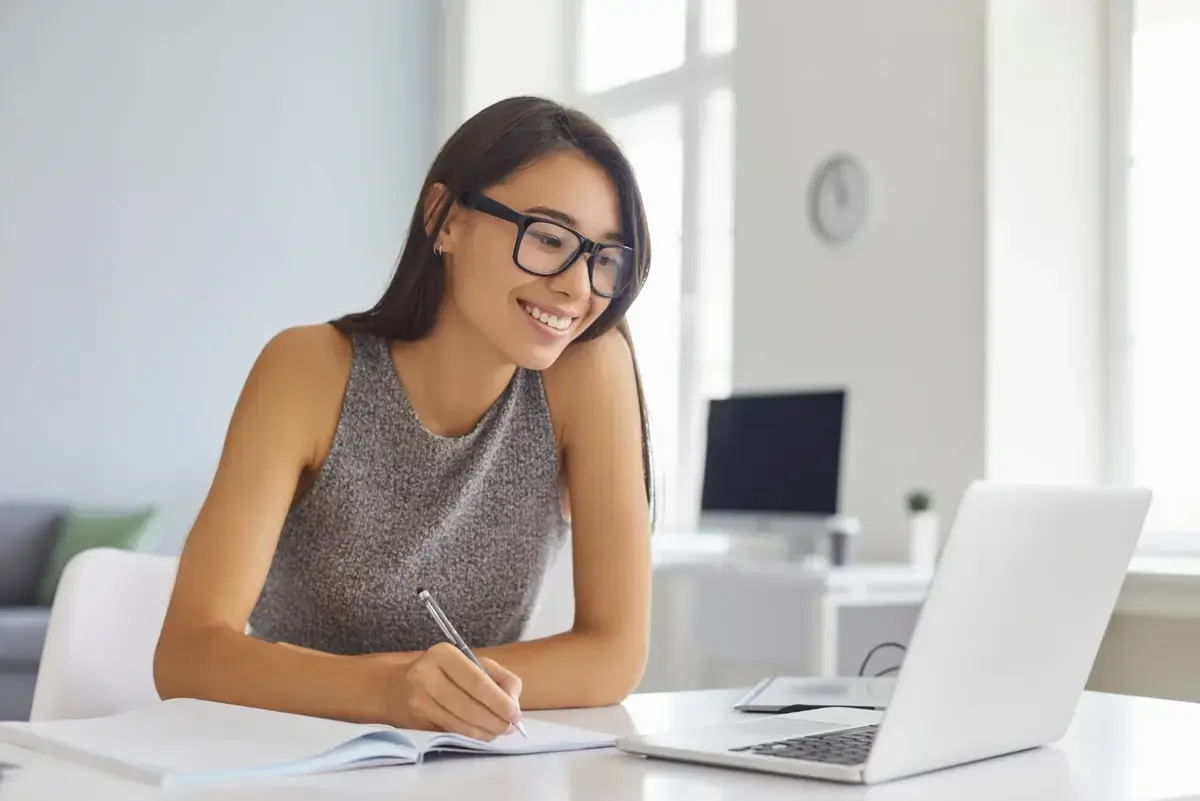 Woman sat working at a laptop with a pen in hand and pad on the table. 